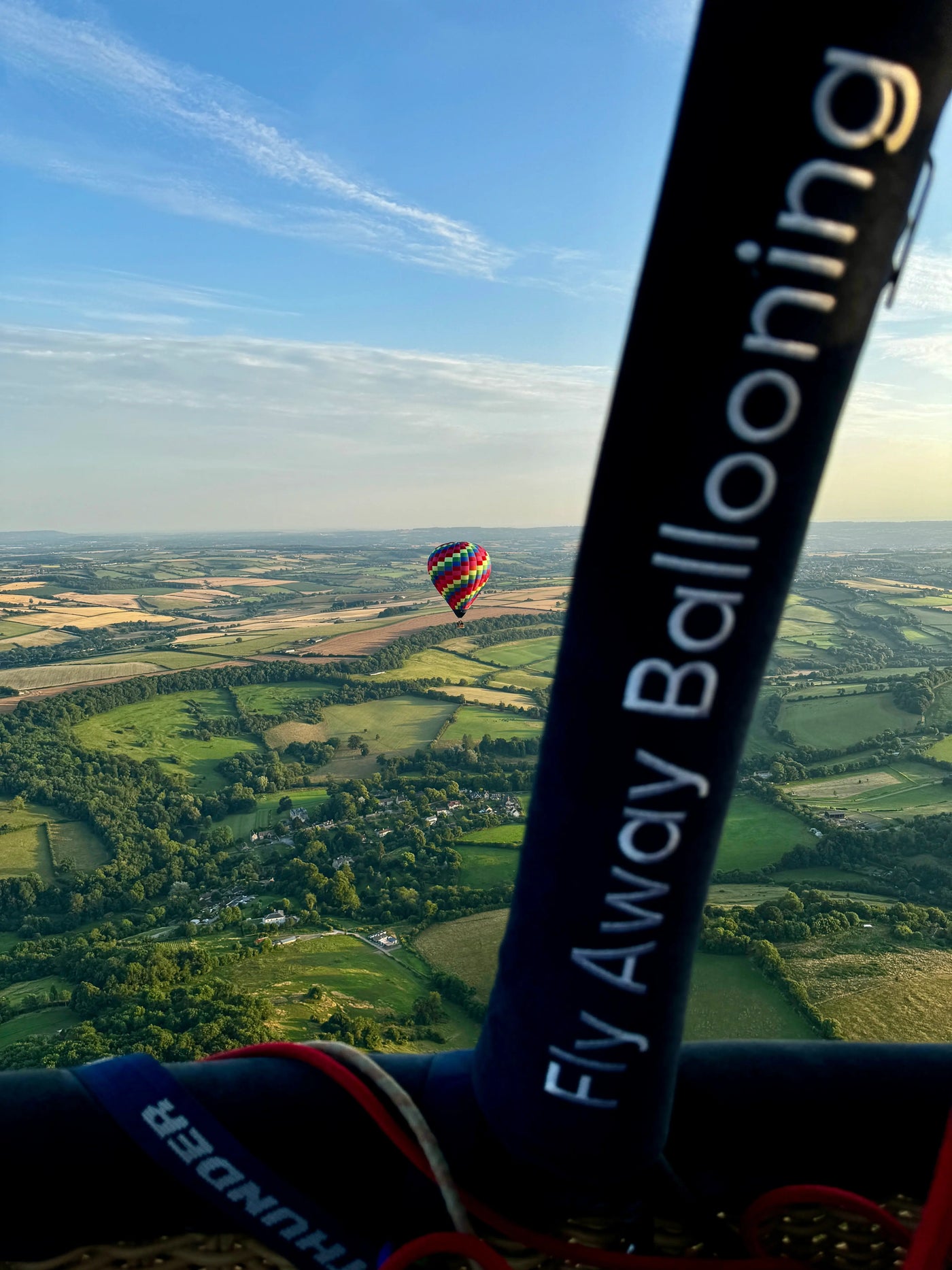 View from a hot air balloon basket looking towards another colourful balloon flying over Somerset countryside