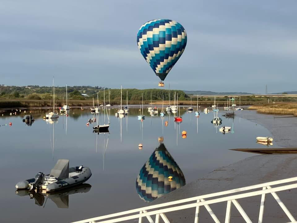 hot air balloon floating over a calm marina at Topsham, Devon with boats and reflections in the water during a morning balloon ride.