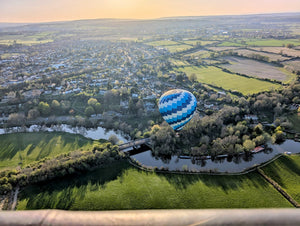 Scenic hot air balloon rides over South West England countryside at sunset, with panoramic views of green fields, a winding river, and a peaceful village below.