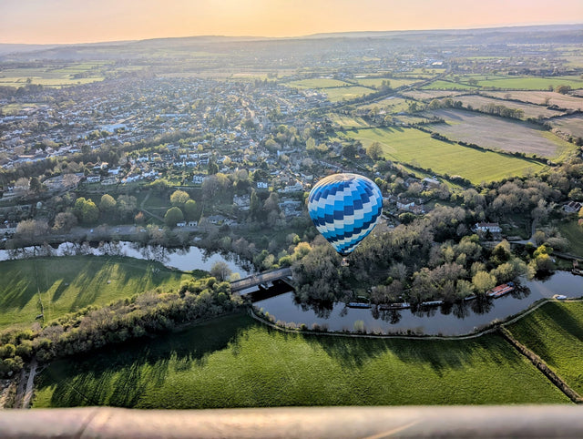 Scenic hot air balloon rides over South West England countryside at sunset, with panoramic views of green fields, a winding river, and a peaceful village below.